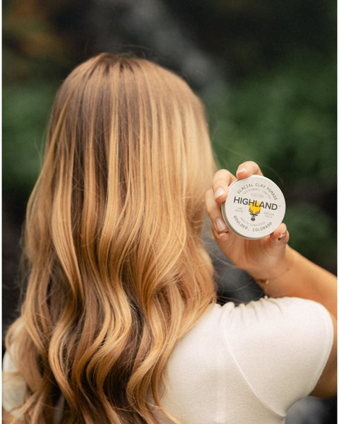 Person with long wavy blonde hair holding a tin of Highland Glacial Clay Pomade against a blurred green background