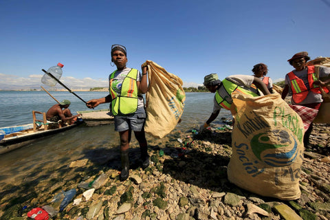 Volunteers collecting plastic waste along a shoreline, filling large sacks with trash while a small boat waits in the water