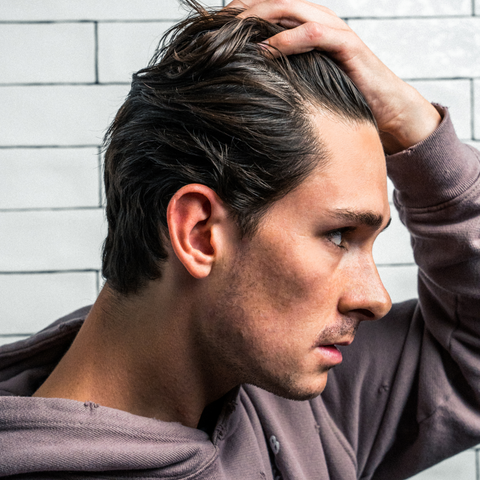 Side profile of a man slicking back pomaded hair against a white subway-tile bathroom wall.