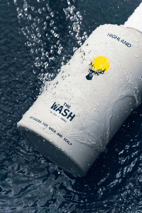 Angled close-up of The Wash bottle showing the THE WASH label and 300ml marking, covered in running water droplets on a dark wet background