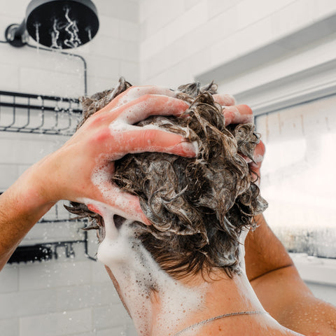 Hands massaging shampoo into wet hair under a showerhead, thick lather covering scalp and neck