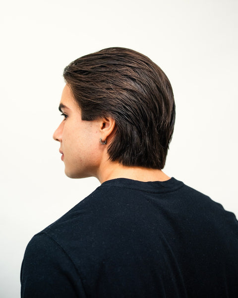 Young man in three-quarter back profile with dark slicked-back hair, wearing a navy sweater on a white background.