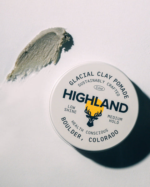 Top-down view of Glacial Clay Pomade tin with Highland branding, 2.1 oz and low shine medium hold, with a smear of product on white