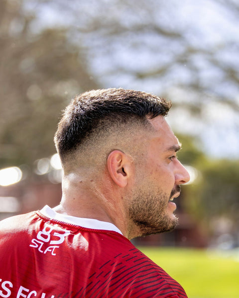 Man in profile showing an Ivy League haircut with a low skin fade, outdoors with blurred trees in the background