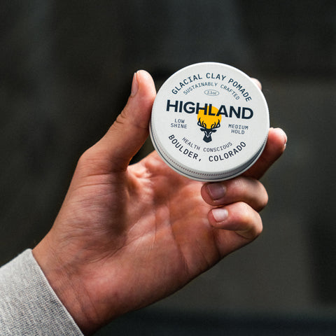 Close-up of hand holding Highland Glacial Clay Pomade tin showing logo and low shine medium hold label against a blurred dark background