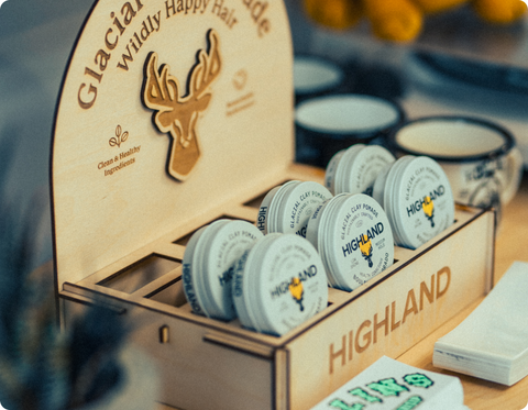 Wooden display box holding small round product tins on a counter; HIGHLAND is printed on the box front.