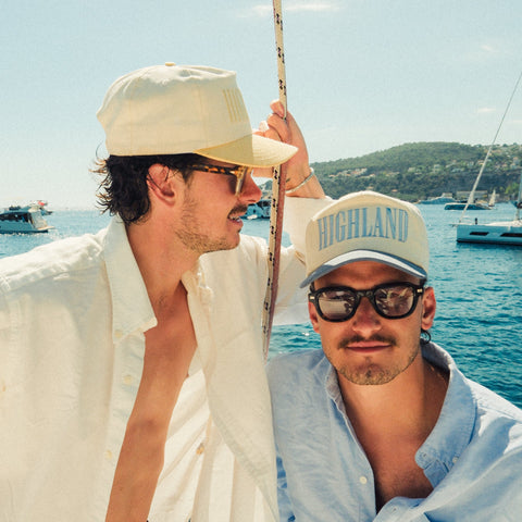 Two men on a sunlit boat wearing caps and sunglasses, yachts and hills on the horizon.