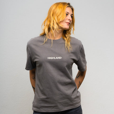 Woman wearing gray Highland Essential Tee with 'HIGHLAND' chest logo, hair blond, hands behind back
