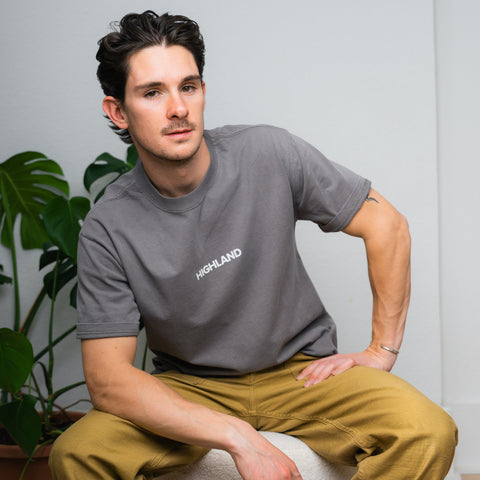 Young man seated on a stool wearing a gray t-shirt and khaki pants, looking to his left.