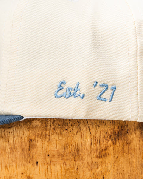 Cream baseball cap close-up showing blue embroidered text: Est. '21