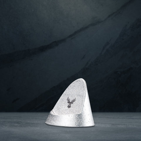 Silver triangular pomade stand with stamped Highland logo on front, on a dark slate surface