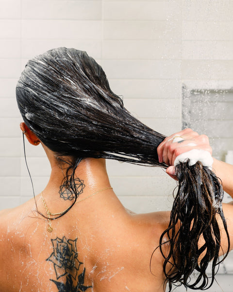 Back view of person shampooing long dark hair, lathered and holding ponytail; neck mandala and rose tattoos, gold necklace.