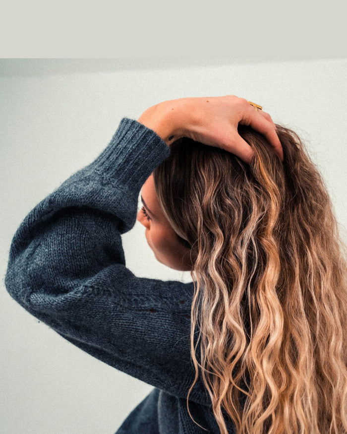 Person lifting a hand through long wavy highlighted hair against a plain light background