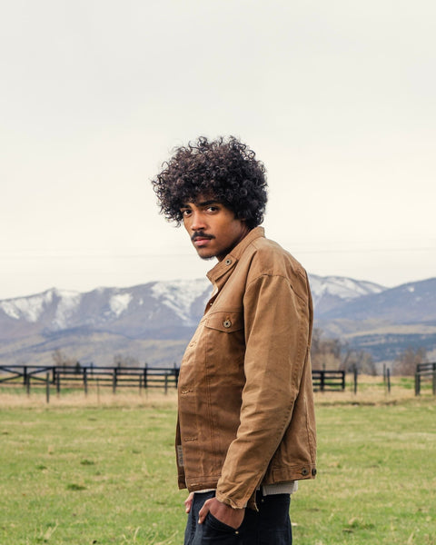 Man with coarse curly hair looking over his shoulder in a field with snow-capped mountains in the background