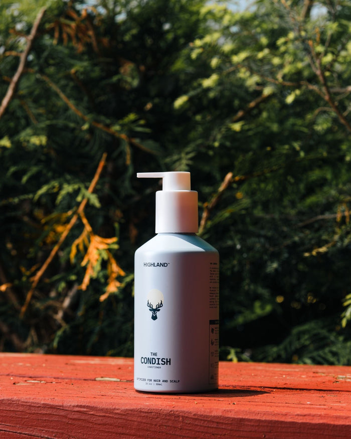 Front view of The Condish conditioner bottle with pump and Highland logo on red wood, blurred green foliage behind