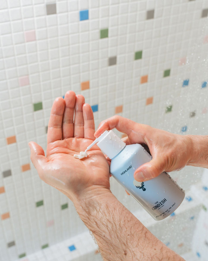 Close-up of The Condish conditioner bottle dispensing cream into wet hands, showcasing pump top and creamy texture with tiled shower background