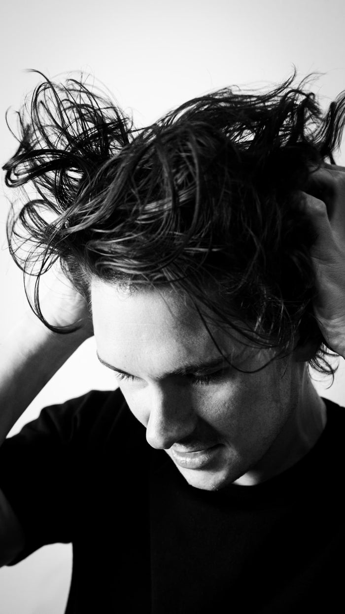 Black-and-white portrait of a person pulling their hair back with both hands, looking down.