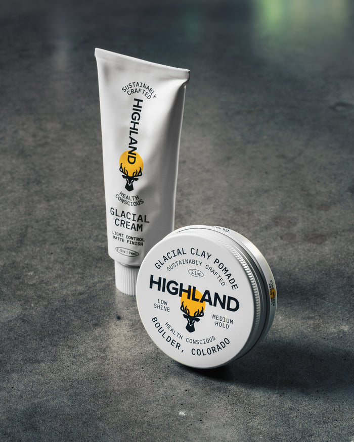 Two Highland grooming products on concrete: Glacial Cream tube and Glacial Clay Pomade tin with Highland logo and yellow sun.