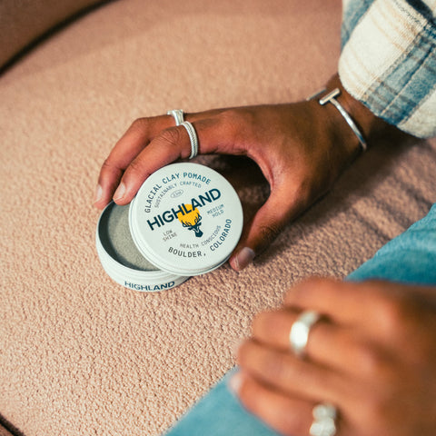 Close-up of hands holding Highland Glacial Clay Pomade tin, label visible and open metal lid on textured beige carpet