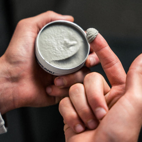 Open tin with gray pomade, hand holding tin and fingertip scooping product