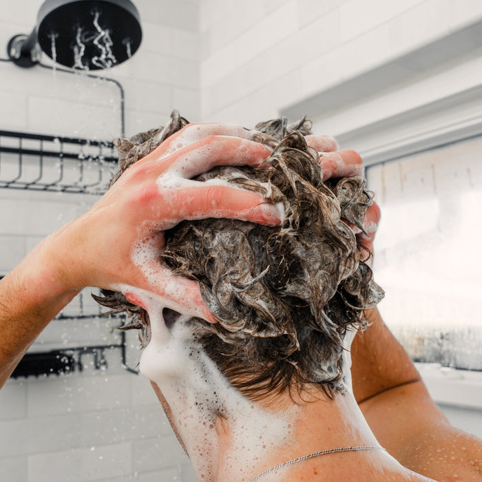 Hands massaging shampoo into wet hair under a showerhead, thick lather covering scalp and neck