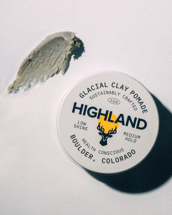 Top-down view of Glacial Clay Pomade tin with Highland branding, 2.1 oz and low shine medium hold, with a smear of product on white