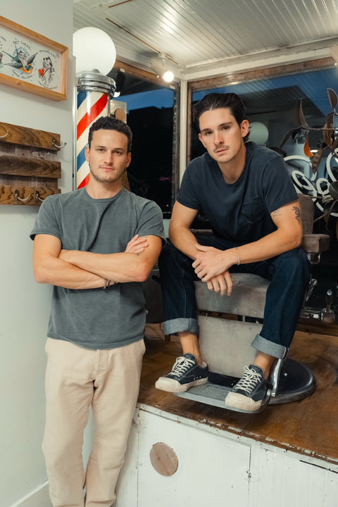 Two men in a barbershop: one seated in a chair, one standing with arms crossed by a barber pole. GET THERE WHILE YOU CAN