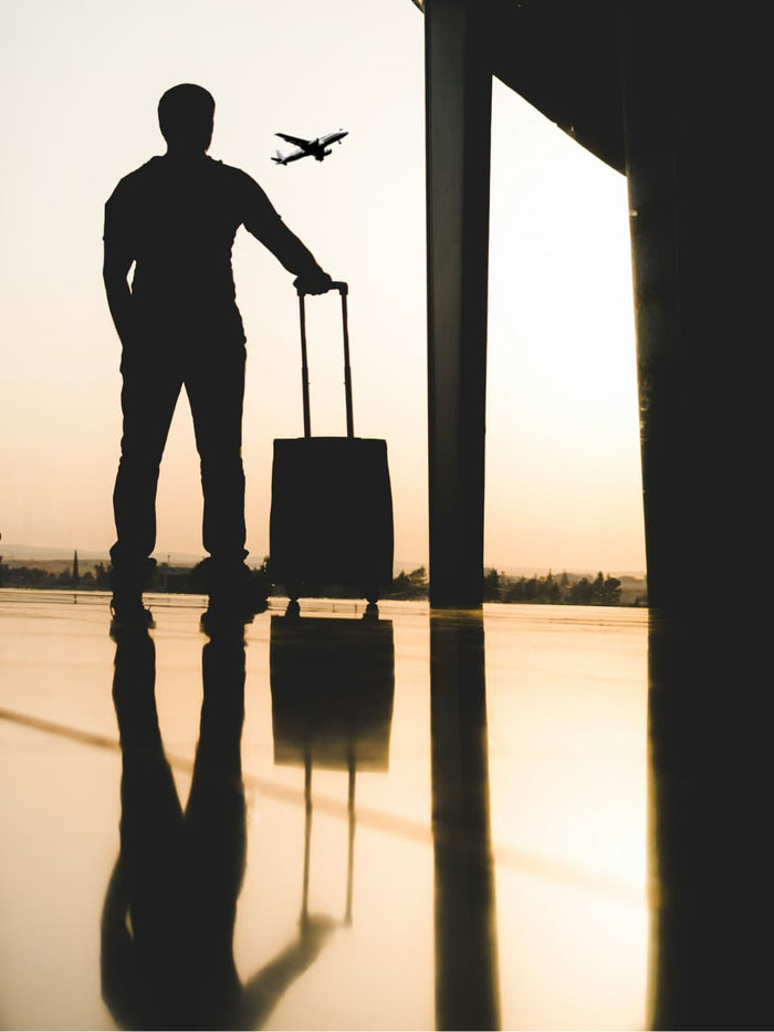 Silhouette of a traveler standing with a carry-on suitcase watching a plane take off, reflected on shiny airport floor
