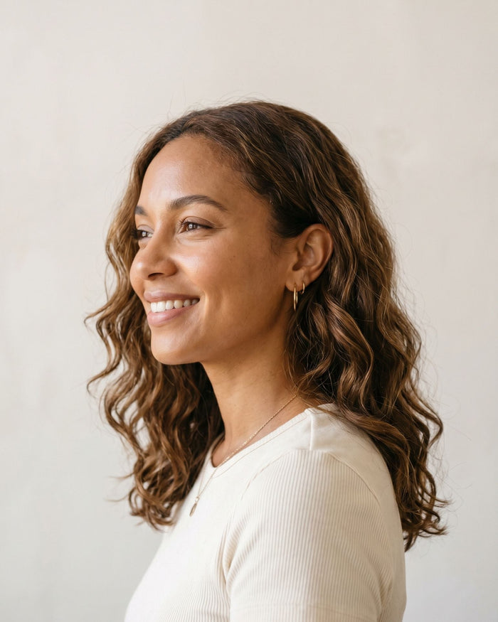 Happy woman with medium-brown skin and soft, defined wavy hair looking away from the camera against a light neutral background.