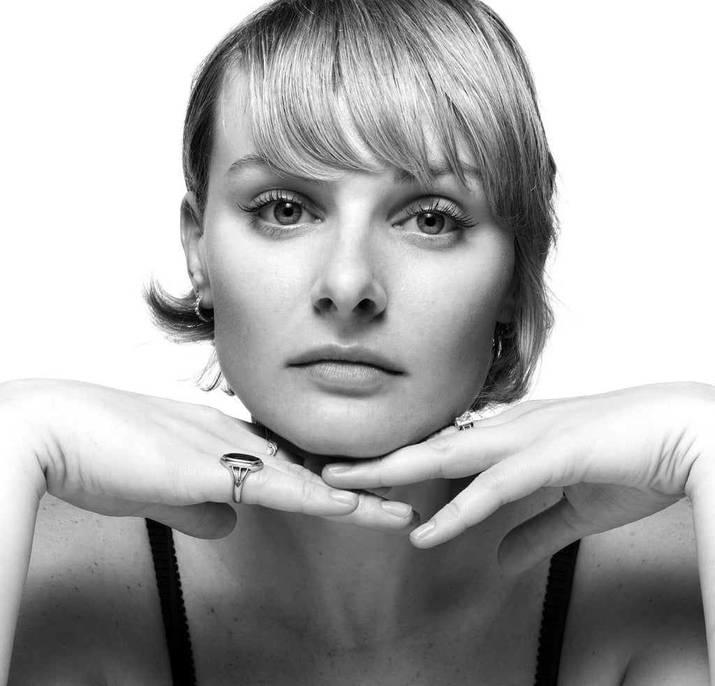 Black-and-white close-up of a young woman resting her chin on crossed hands, neutral expression, short bangs