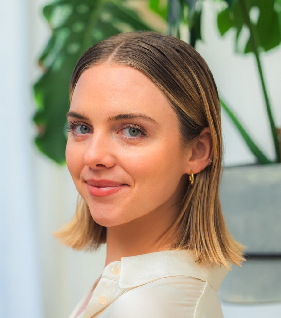 Smiling young woman with shoulder-length blonde hair, white collared shirt, gold hoop earring, soft indoor-plant background.