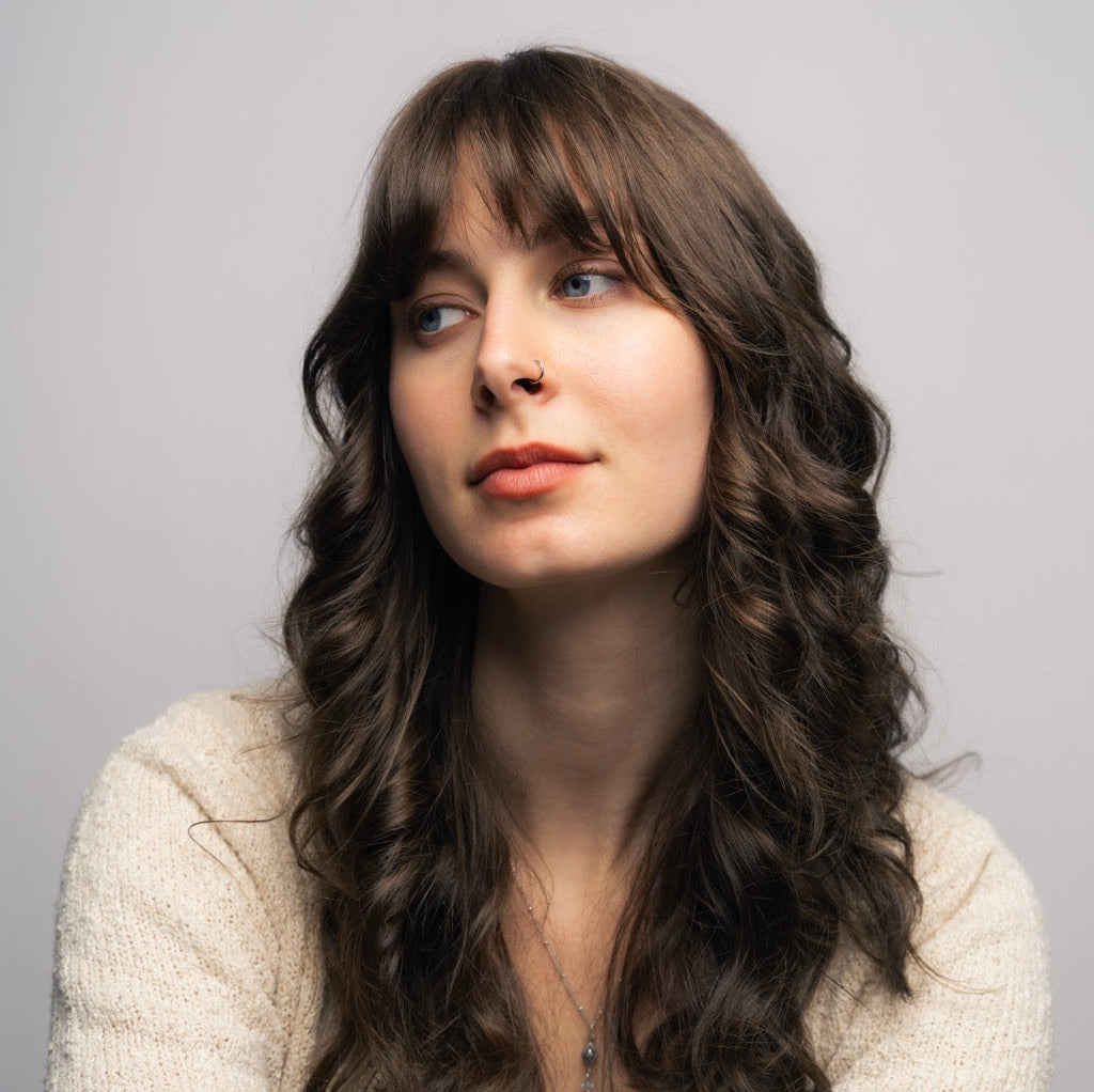 Young woman with long wavy brown hair, nose ring, wearing a light sweater, looking to her right.