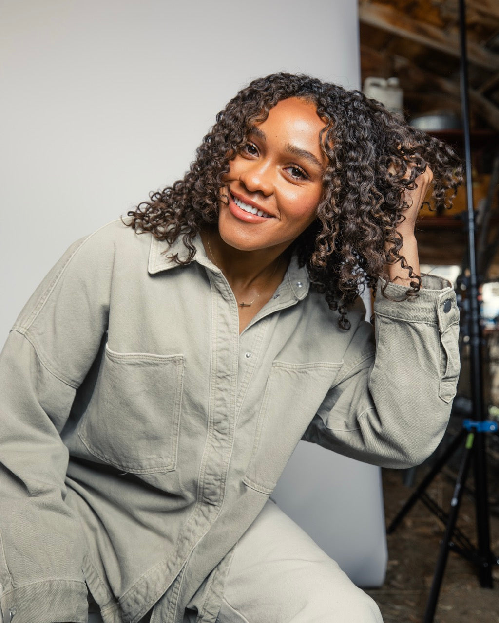 Smiling woman with curly hair in a light gray work shirt, holding hair back in a studio with backdrop and light stand.