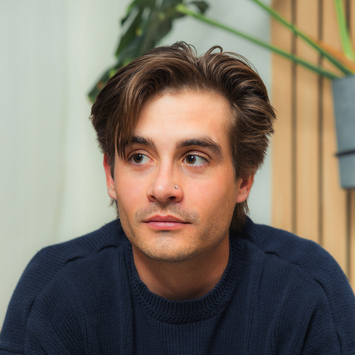 Young man with dark hair in navy sweater looking to the right, indoor plant and wooden slat wall behind him.