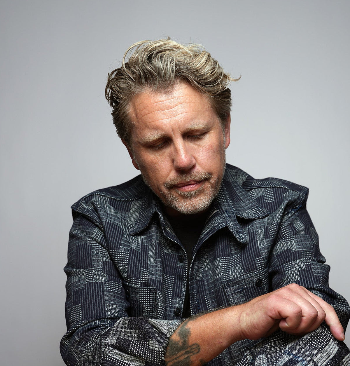 Middle-aged man with gray hair and stubble, in a patterned jacket, looking down with folded arms and forearm tattoo.