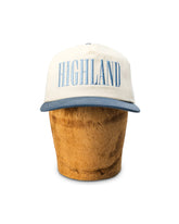 White and blue trucker cap with HIGHLAND on front, displayed on a cylindrical brown hat block