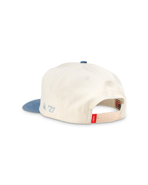 Cream baseball cap with blue brim, rear view showing adjustable strap and small red tag.