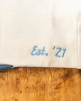 Cream baseball cap close-up showing blue embroidered text: Est. &#39;21