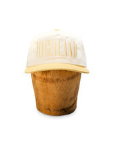 Cream baseball cap with raised HIGHLAND lettering on a brown cylindrical display stand.