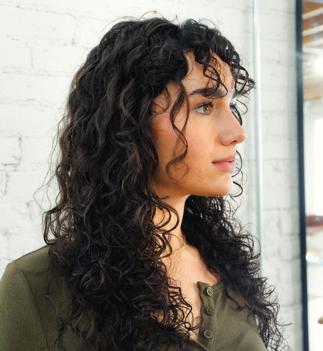 Young woman with curly dark hair in profile looking right, standing by a white brick wall and window reflection.