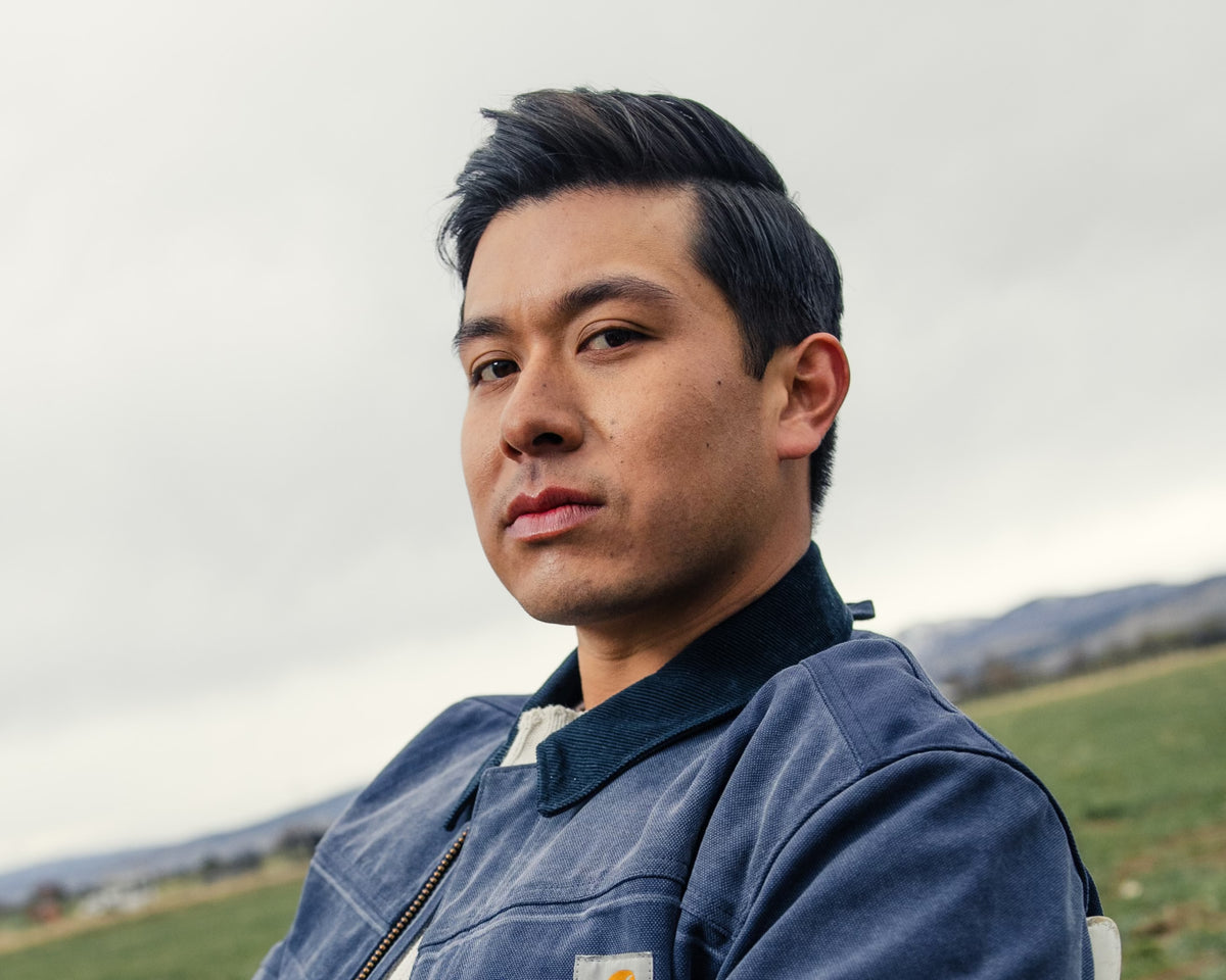 Man in a blue work jacket standing outdoors in a field under an overcast sky, looking at camera.