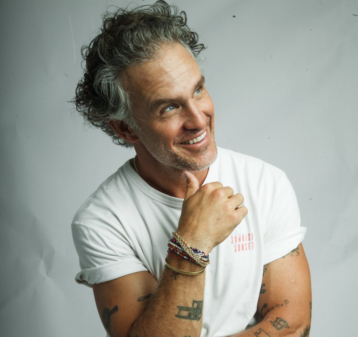 Smiling gray-haired man with tattoos and bracelets, wearing a white T-shirt reading 'SUNRISE SUNSET', looking up and to the right.