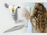Tube of hair treatment, a gray product smear, and a woman with long curly hair looking into a mirror