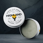 Highland Glacial Clay Pomade 2.1 oz tin, open with the lid propped beside it, showing the grey-green clay pomade inside. The lid reads low shine, medium hold.