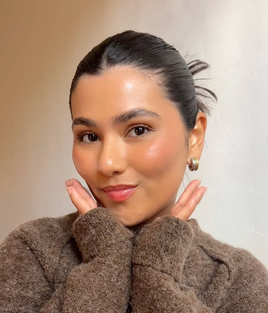 Woman with slicked-back bun, gold hoop earrings, brown fuzzy sweater, hands framing her smiling face.