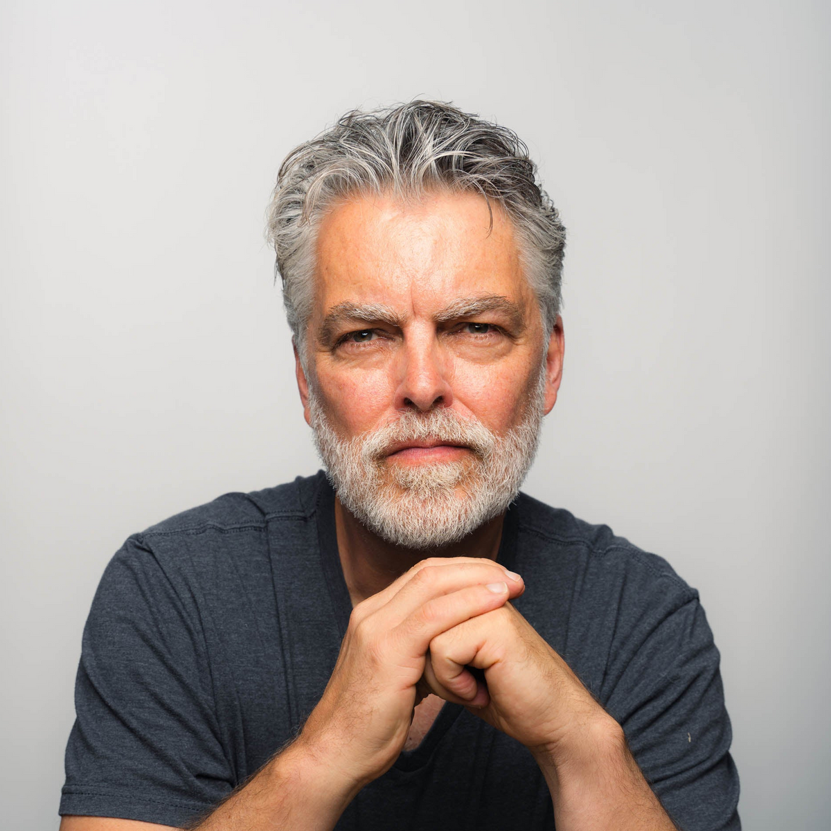Portrait of serious older man with gray hair and beard, hands clasped, wearing dark T-shirt on light background.