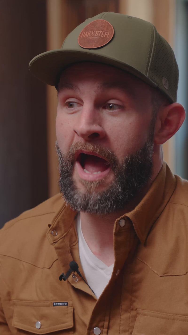 Bearded man speaking, mouth open, wearing green cap with patch OAK&STEEL and brown shirt labeled PONCHO, clip-on mic visible.