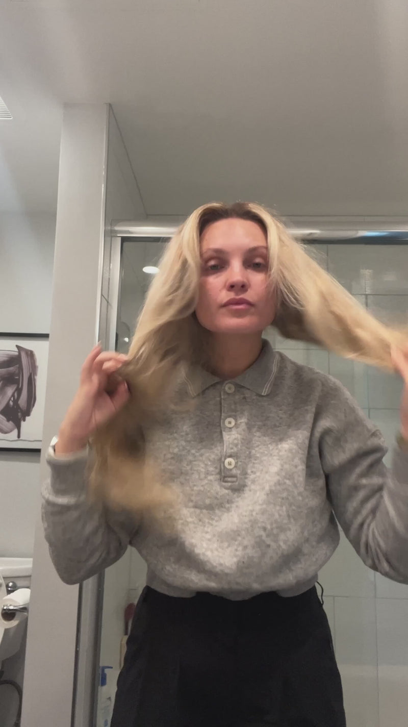 Person with long blonde hair holding hair to show frizz in a bathroom with a glass shower door.