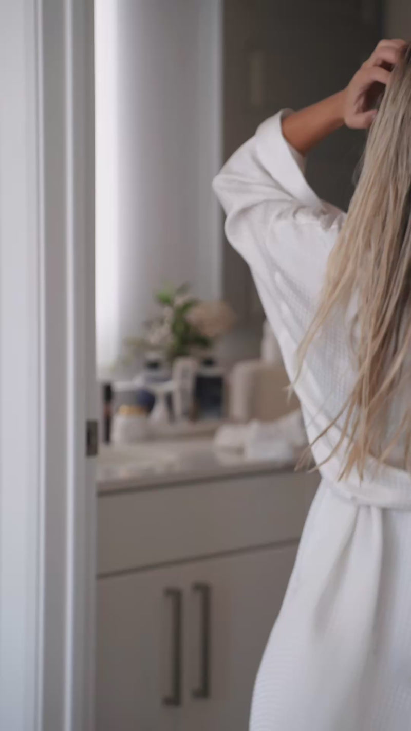 Back view of person in white robe running hand through long damp hair by a bathroom vanity.