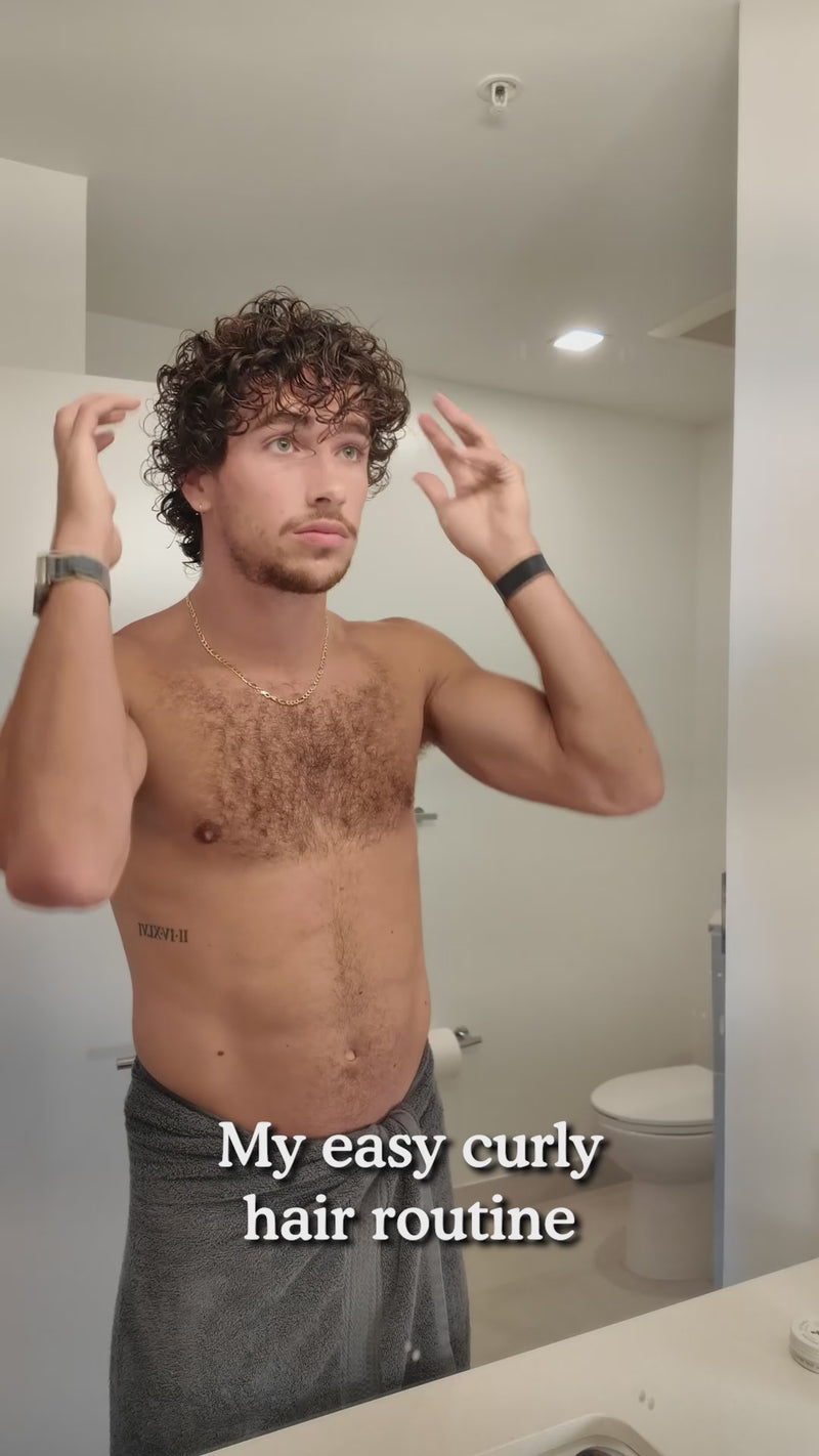 Man adjusting curly hair in bathroom mirror with towel at waist; overlay text: My easy curly hair routine